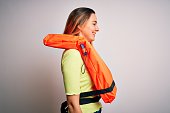 Young beautiful blonde woman with blue eyes wearing orange lifejacket over white background looking to side, relax profile pose with natural face with confident smile.