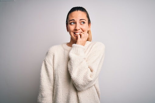 Young beautiful blonde woman with blue eyes wearing casual sweater over white background looking stressed and nervous with hands on mouth biting nails. Anxiety problem.