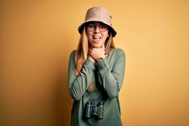 istock Beautiful blonde explorer woman with blue eyes wearing hat and glasses using binoculars shouting and suffocate because painful strangle. Health problem. Asphyxiate and suicide concept. 1212512815