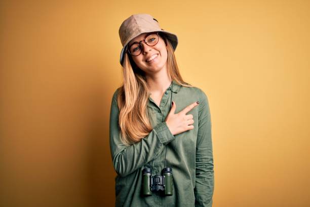 istock Beautiful blonde explorer woman with blue eyes wearing hat and glasses using binoculars cheerful with a smile of face pointing with hand and finger up to the side with happy and natural expression on face 1212512817