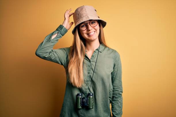 Beautiful blonde explorer woman with blue eyes wearing hat and glasses using binoculars confuse and wonder about question. Uncertain with doubt, thinking with hand on head. Pensive concept.