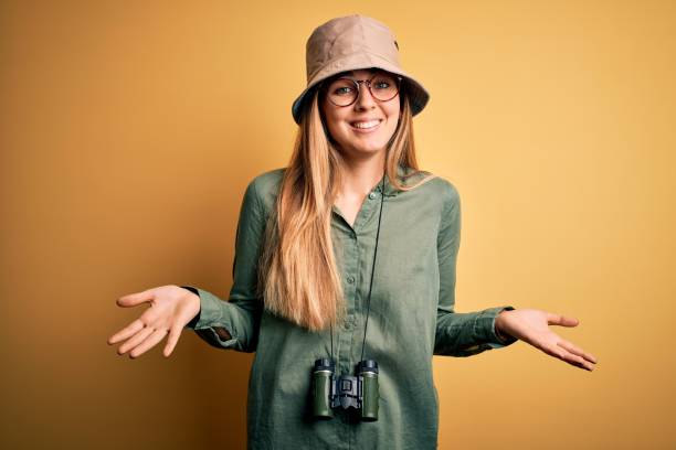 istock Beautiful blonde explorer woman with blue eyes wearing hat and glasses using binoculars clueless and confused expression with arms and hands raised. Doubt concept. 1212512833