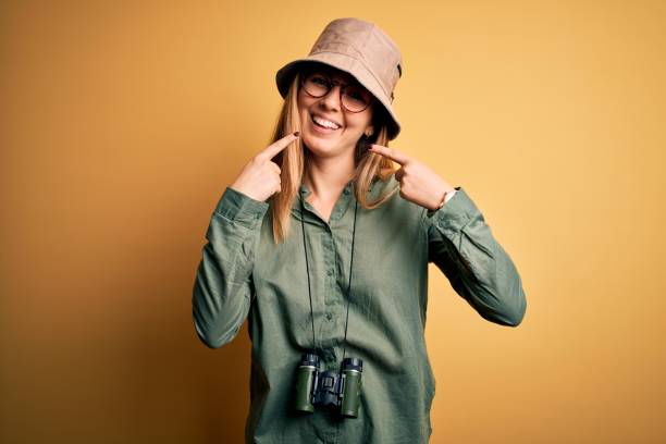 istock Beautiful blonde explorer woman with blue eyes wearing hat and glasses using binoculars smiling cheerful showing and pointing with fingers teeth and mouth. Dental health concept. 1212512835