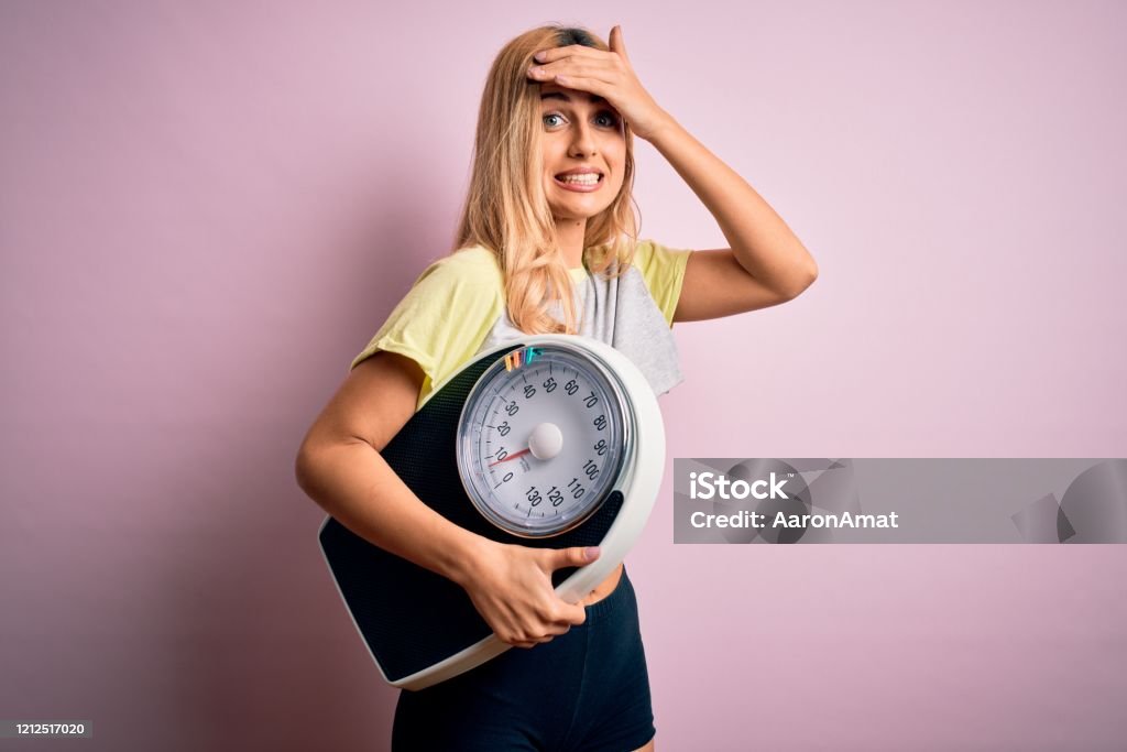 Young beautiful blonde sporty woman on diet holding weight machine over pink background stressed with hand on head, shocked with shame and surprise face, angry and frustrated. Fear and upset for mistake. Dieting Stock Photo