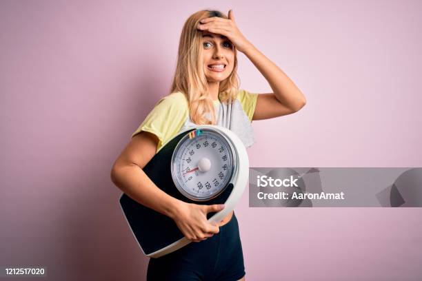 Young Beautiful Blonde Sporty Woman On Diet Holding Weight Machine Over Pink Background Stressed With Hand On Head Shocked With Shame And Surprise Face Angry And Frustrated Fear And Upset For Mistake Stock Photo - Download Image Now