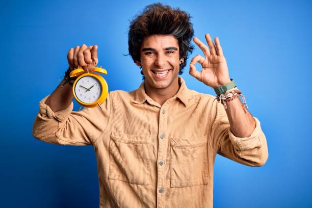 istock Young handsome man holding alarm clock standing over isolated blue background doing ok sign with fingers, excellent symbol 1212517984