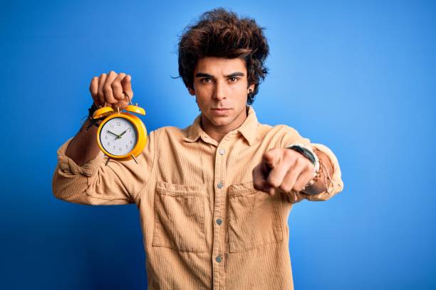 istock Young handsome man holding alarm clock standing over isolated blue background pointing with finger to the camera and to you, hand sign, positive and confident gesture from the front 1212517993