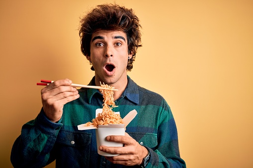 Young handsome man eating noodles standing over isolated yellow background scared in shock with a surprise face, afraid and excited with fear expression