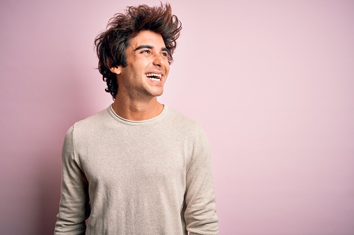Young handsome man wearing casual t-shirt standing over isolated pink background looking away to side with smile on face, natural expression. Laughing confident.