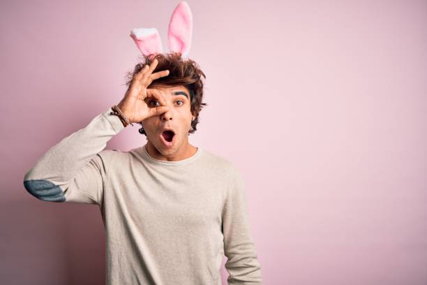 istock Young handsome man holding easter rabbit ears standing over isolated pink background doing ok gesture shocked with surprised face, eye looking through fingers. Unbelieving expression. 1212518945