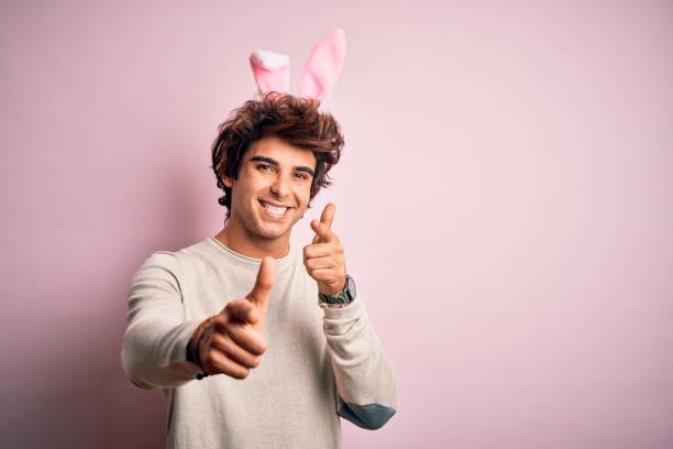 Amazed young Caucasian man in bunny ears isolated on pink studio background point at blank copy space. Stunned male in rabbit costume shocked by good sale offer or promotion deal.