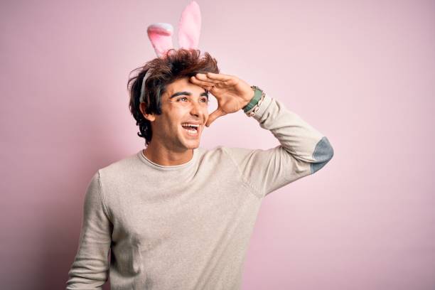 istock Young handsome man holding easter rabbit ears standing over isolated pink background very happy and smiling looking far away with hand over head. Searching concept. 1212518990
