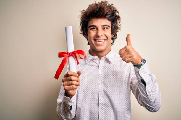 Young handsome student man holding diploma standing over isolated pink background happy with big smile doing ok sign, thumb up with fingers, excellent sign
