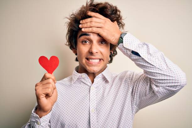 istock Young handsome man holding paper heart standing over isolated white background stressed with hand on head, shocked with shame and surprise face, angry and frustrated. Fear and upset for mistake. 1212519741