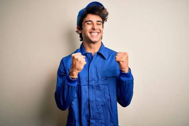 Photo of Young mechanic man wearing blue cap and uniform standing over isolated white background excited for success with arms raised and eyes closed celebrating victory smiling. Winner concept.