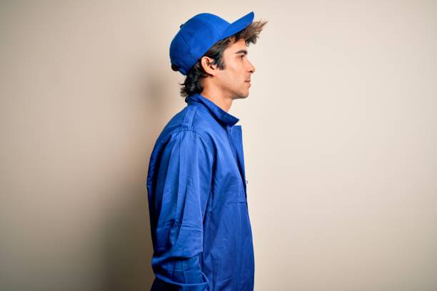 Young mechanic man wearing blue cap and uniform standing over isolated white background looking to side, relax profile pose with natural face with confident smile.