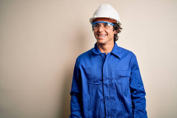 Young constructor man wearing uniform and security helmet over isolated white background looking away to side with smile on face, natural expression. Laughing confident.