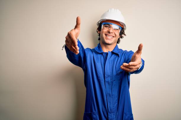 istock Young constructor man wearing uniform and security helmet over isolated white background looking at the camera smiling with open arms for hug. Cheerful expression embracing happiness. 1212519982