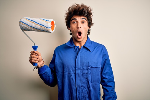 Young handsome painter man holding roller wearing uniform over isolated white background scared in shock with a surprise face, afraid and excited with fear expression