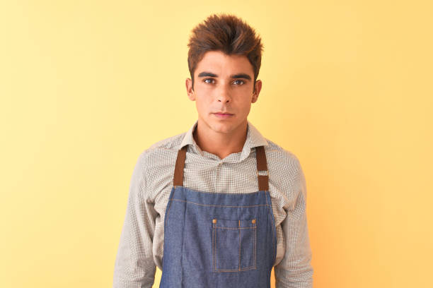 istock Young handsome employee man wearing apron standing over isolated yellow background with serious expression on face. Simple and natural looking at the camera. 1212521297