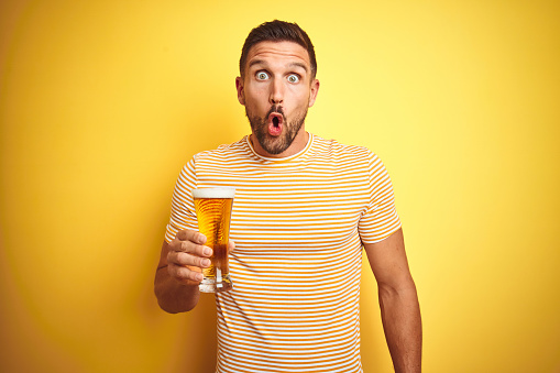 Young handsome man drinking a pint glass of beer over isolated yellow background scared in shock with a surprise face, afraid and excited with fear expression