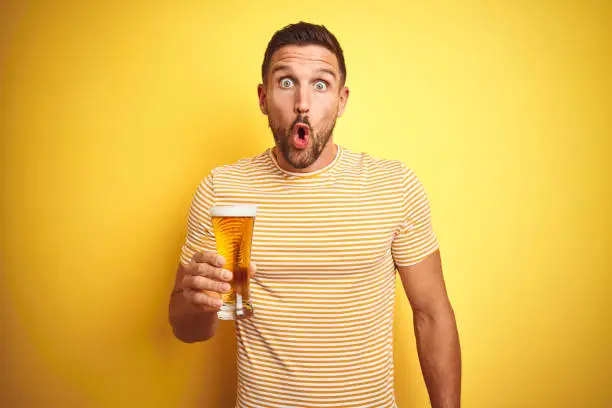 Photo of Young handsome man drinking a pint glass of beer over isolated yellow background scared in shock with a surprise face, afraid and excited with fear expression