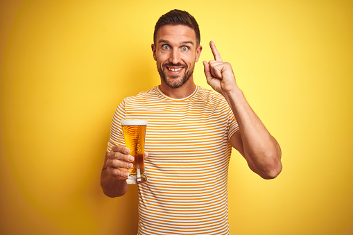 Young handsome man drinking a pint glass of beer over isolated yellow background surprised with an idea or question pointing finger with happy face, number one