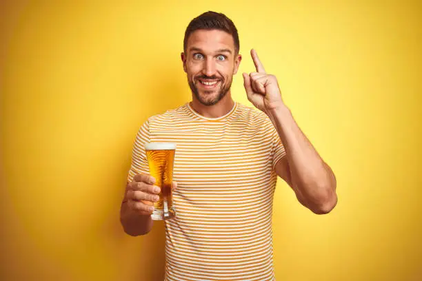 Photo of Young handsome man drinking a pint glass of beer over isolated yellow background surprised with an idea or question pointing finger with happy face, number one