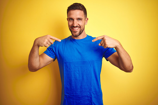 Young handsome man wearing casual blue t-shirt over yellow isolated background looking confident with smile on face, pointing oneself with fingers proud and happy.