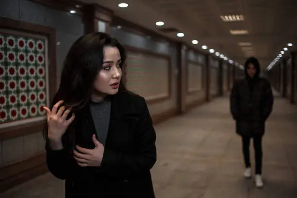 Photo of a young beautiful woman walks at night, followed by a man in dark clothes with a hood on his head. mosaics on the walls are not someone's works of art.these are walls in Underpass in an Oriental style