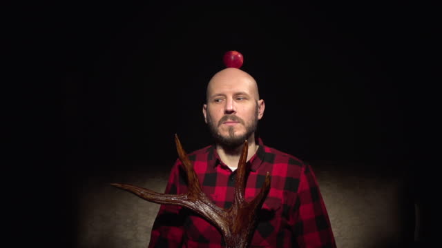 Mature eyes rolling bald caucasian white young hipster man with beard in red plaid shirt with moose deer horns in hands red apple on top of head