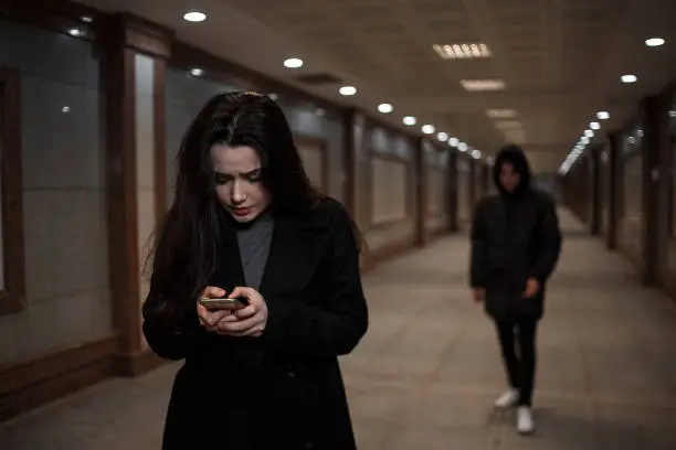 Photo of a young beautiful woman followed by a man in dark clothing with a hood on his head. a woman tries to dial the number of the rescue service on the phone. mosaics on the walls are not paintings