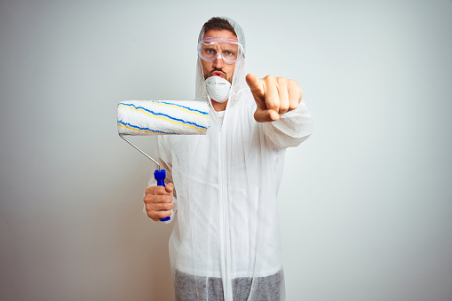 Painter man wearing professional worker equipment holding roller over isolated background pointing with finger to the camera and to you, hand sign, positive and confident gesture from the front