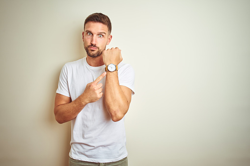 Young handsome man wearing casual white t-shirt over isolated background In hurry pointing to watch time, impatience, looking at the camera with relaxed expression