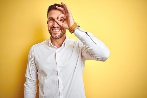 Young handsome man wearing elegant white shirt over yellow isolated background doing ok gesture with hand smiling, eye looking through fingers with happy face.