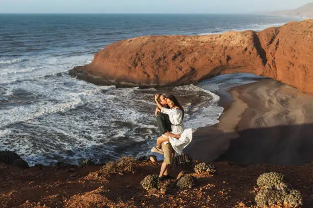 Photo of Young happy smiling couple hugging in love on background of famous arch on Legzira beach in Morocco. Sidi Ifni landscape. Popular moroccan landmark.
