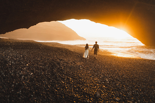 Couple walking under big arch on Legzira ocean beach and enjoying fantastic warm sunset. View from back. Famous and popular landmark in Morocco. Beautiful evening light.