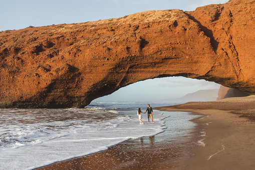 Couple in love walking under huge arch on Legzira ocean beach at sunset. Famous and popular landmark in Morocco.