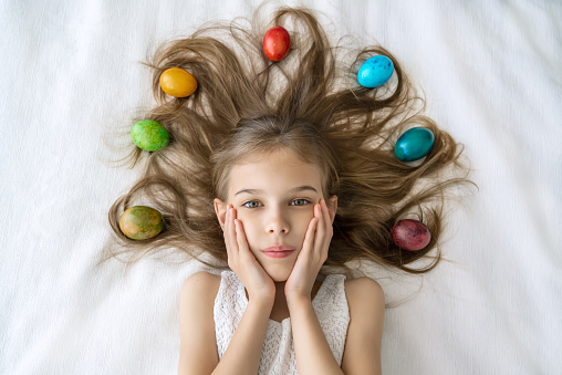 Close up portrait of a little girl, holding hands on her face and decoration of colorful easter eggs in her long hair