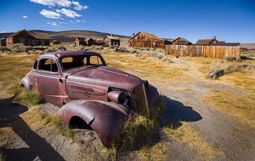Rusty oldtimer in ghost town Bodie, an old gold digger town in California, wooden houses in the background, blue sunny sky, USA