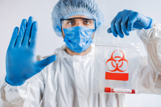 istock Scientist in biological protective costume showing stop gesture with biohazard trasportation package. Epidemic virus concept 1212567737