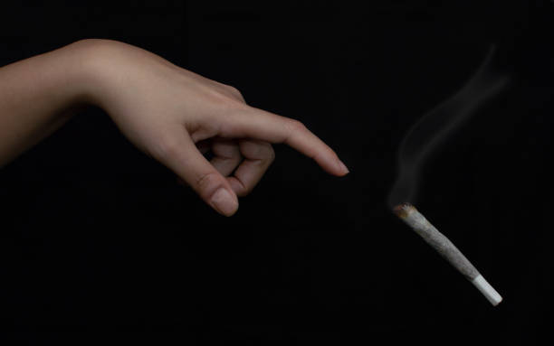 Artistic cannabis background: Hand about to touch a big lit joint of marihuana or cigarette isolated on black. Concept of addiction or start smoking. Artistic cannabis background: Hand about to touch a big lit joint of marihuana or cigarette isolated on black. Concept of addiction or start smoking. pitter stock pictures, royalty-free photos & images
