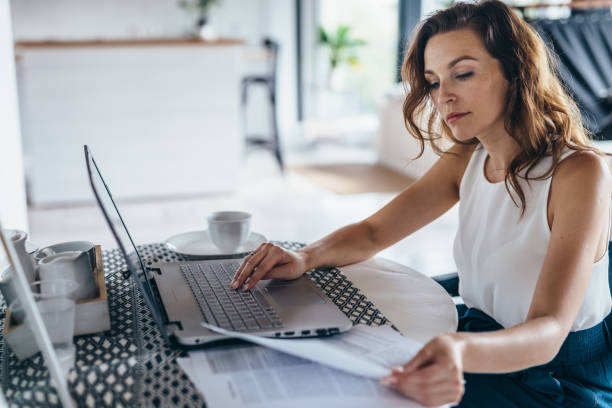 femme utilisant l’ordinateur portatif tout en s’asseyant à la table. jeune femme d’affaires s’asseyant dans la cuisine et travaillant sur l’ordinateur portatif. - home worker photos et images de collection