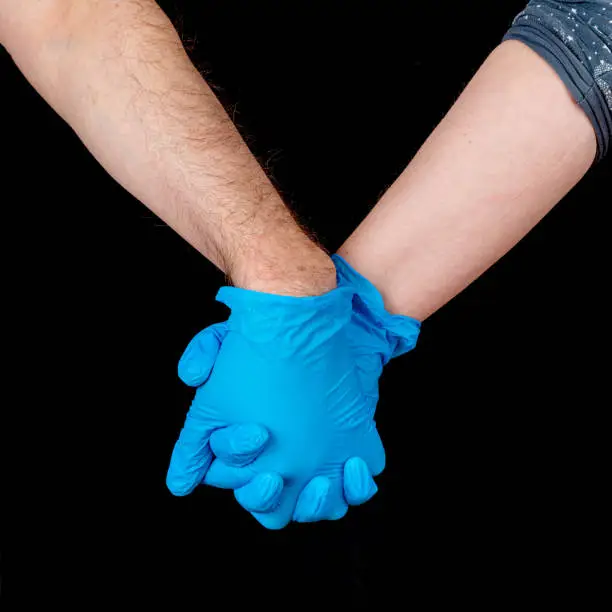 A couple hold hands while wearing blue latex gloves. Closeup view showing only the hands. Black background.
