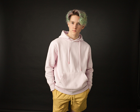 studio closeup portrait of a teenager boy with green hair in a pink hooded shirt and yellow shorts on a black background
