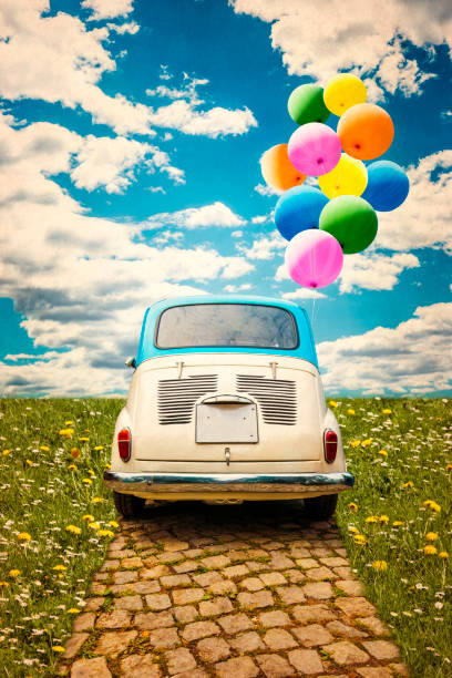 Vintage car with colorful balloons riding through pavement between sunny meadow.