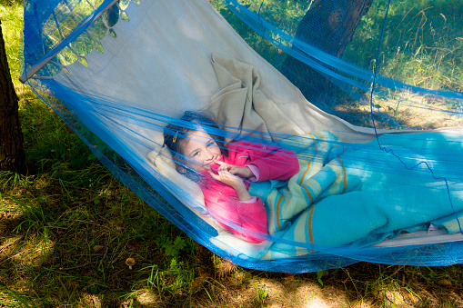 Child girl relaxing in a sunny hammock with mosquito net in forest.