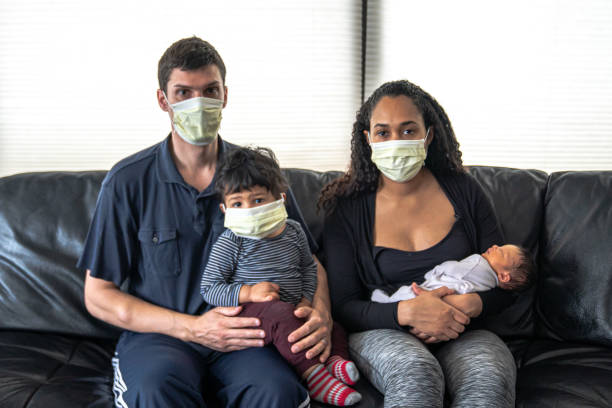 a mixed family sits on a black leather sofa with father and mother holding their infant son and daughter all wearing medical face masks in hopes of preventing getting sick from caronavirus or covid-19 - mother family baby isolated imagens e fotografias de stock