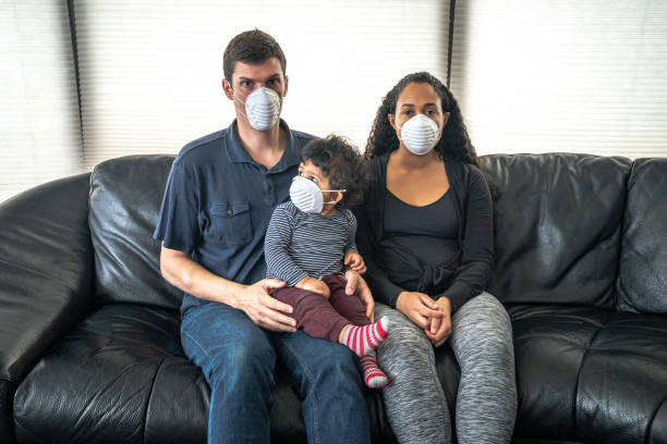 istock A young mixed family sits on a black leather sofa holding their son and wearing a dust face mask over their faces in hopes of preventing getting sick or ill from caronavirus or COVID-19. 1212628024