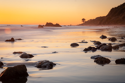 The bright orange sun reflects in the sand and the surf as it sets over the horizon of the Pacific Ocean. The coastal cliffs of Malibu descend into the waves decorated with silhouetted palm trees. Rocky boulders dot the foreground as the surf moves around them and reflects the orange and turquoise colors of the sky. A long exposure makes the water soft and smooth. There are no clouds in the sky, and no people on the beach.